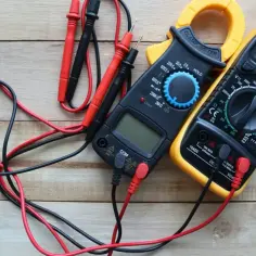 Dwa multimetry cyfrowe, jeden czarny z żółtą obudową, drugi czarny, leżą na drewnianym tle.